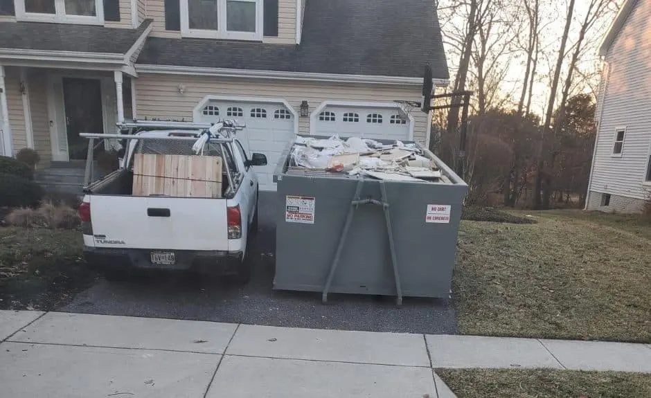 Gray roll-off dumpster filled with renovation debris in a home driveway next to a white contractor pickup truck.