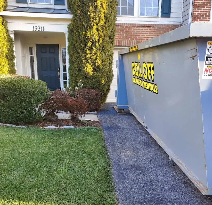 Gray Roll Off On The Go Rentals roll-off dumpster placed on a driveway beside the front walk of a brick house.