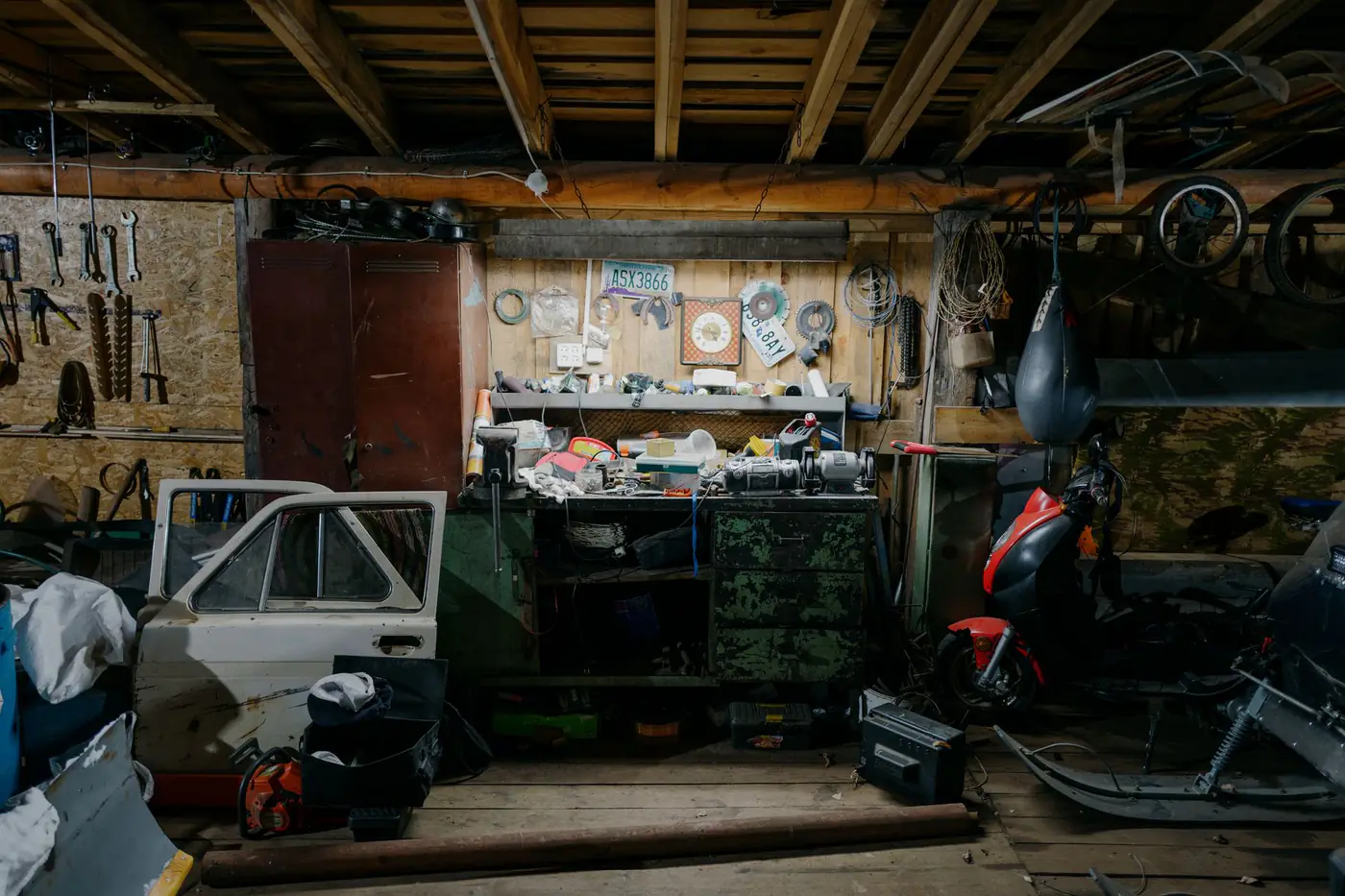 Crowded garage workshop packed with tools, an old car door, a scooter and piles of clutter