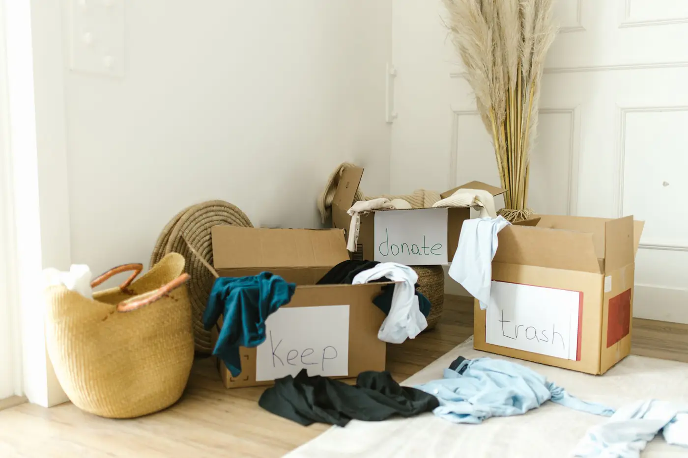 Cardboard boxes labeled keep, donate and trash filled with clothes on a bedroom floor
