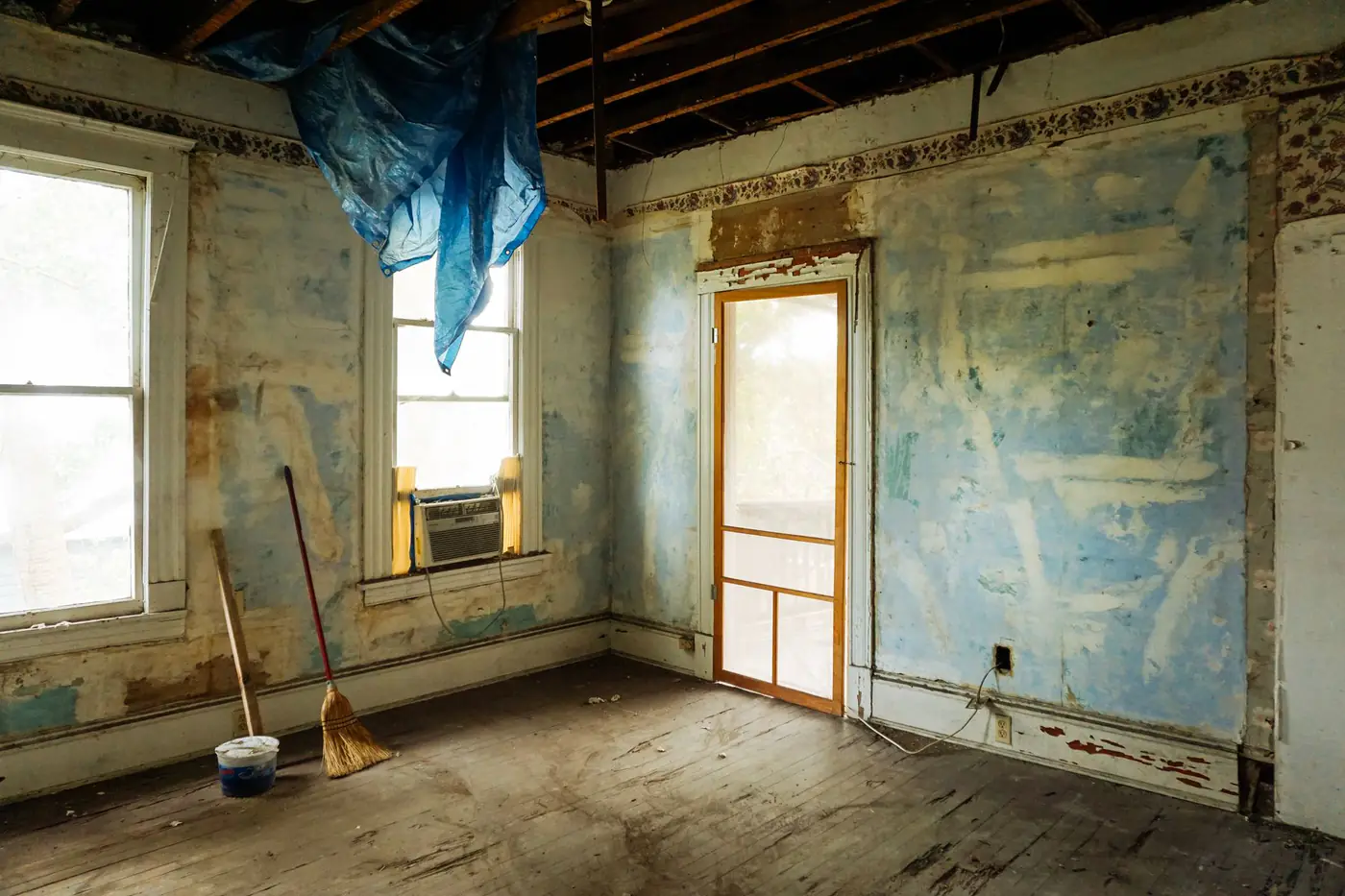 Old room stripped for renovation with exposed ceiling joists, peeling walls, a broom and a bucket on the floor