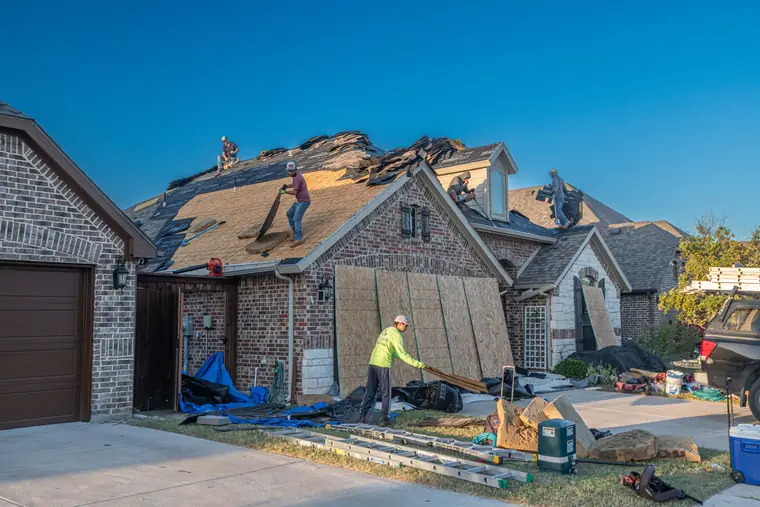 Roofers tearing off old shingles on a brick home while a worker collects debris near the garage and driveway