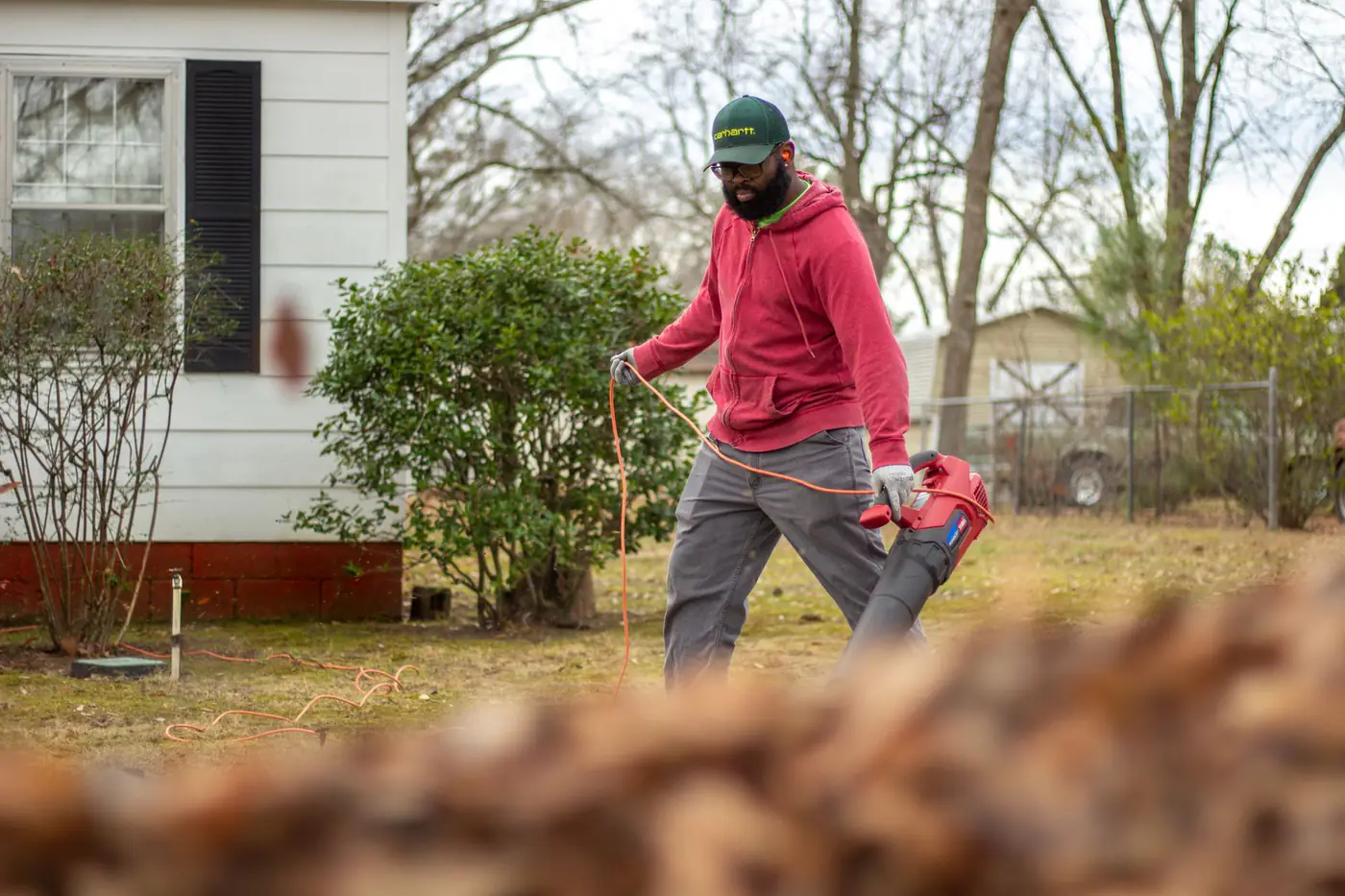 Man in a red hoodie using a leaf blower in the backyard of a white house in early spring